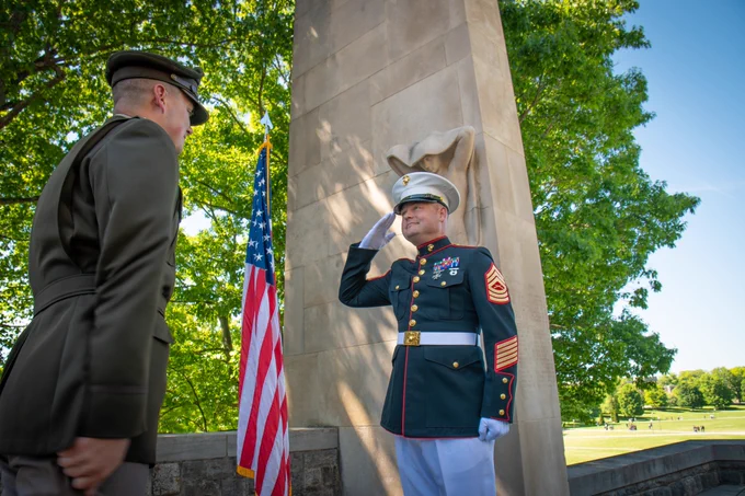 A new officer in a U.S. Army uniform stands at attention while a U.S. Marine in dress uniform renders a salute during a commissioning ceremony beside an American flag and a stone memorial.