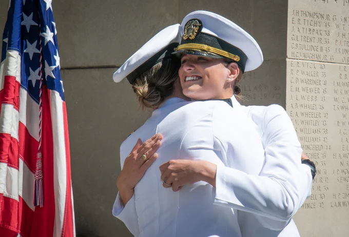 Two commissioned officers in white dress uniforms embrace beside an American flag and a stone memorial engraved with names during a commissioning ceremony.