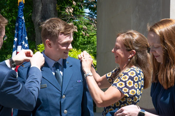Family members adjust shoulder insignia on a newly commissioned officer in a U.S. Air Force dress uniform during a commissioning ceremony beside a stone memorial and American flag.