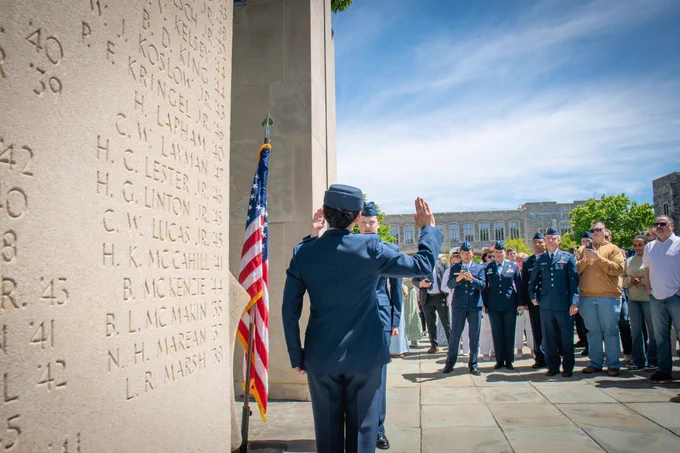 A cadet in a U.S. Air Force dress uniform raises a right hand to take the oath during a commissioning ceremony beside an American flag and a stone memorial, with a crowd gathered in the background.