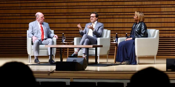 A group of three people in a discussion setting. One person is gesturing while speaking, seated on a stage with two others who are listening attentively.