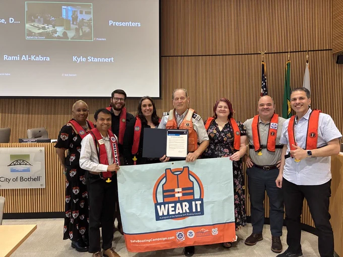A group of people post for a picture in Bothell City Hall Council Chambers. One person in the center holds a proclamation, and a “Wear It” lifeguard poster. The people in the picture are wearing life jackets.
