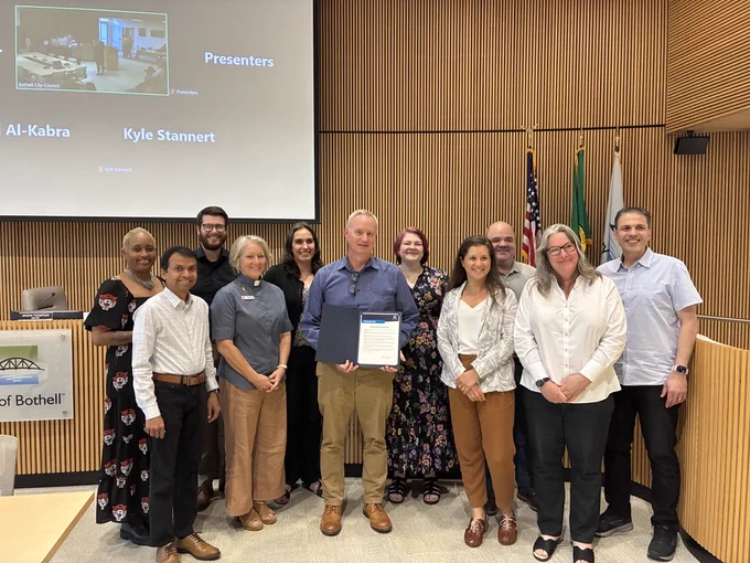 A group of people post for a picture in Bothell City Hall Council Chambers. One person in the center holds a proclamation. 