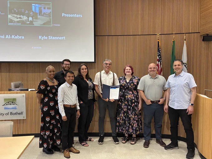 A group of people post for a picture in Bothell City Hall Council Chambers. One person in the center holds a proclamation. 