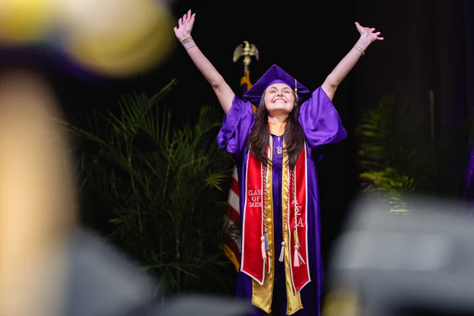 A graduate smiles with their arms raised as they walk across the commencement stage. 