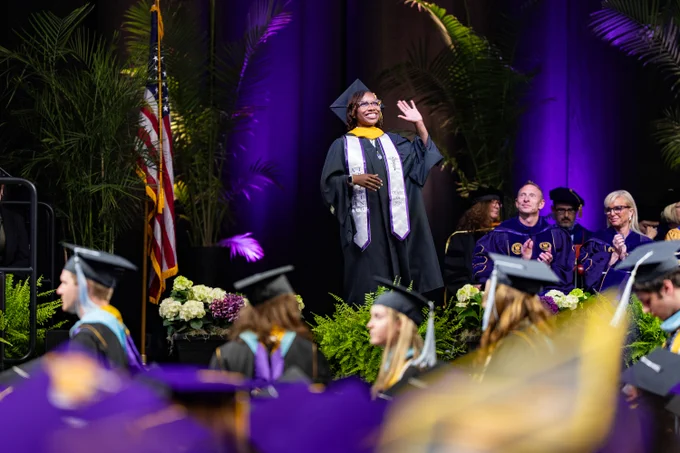 A graduate smiles and waves as they cross the stage during a commencement ceremony.