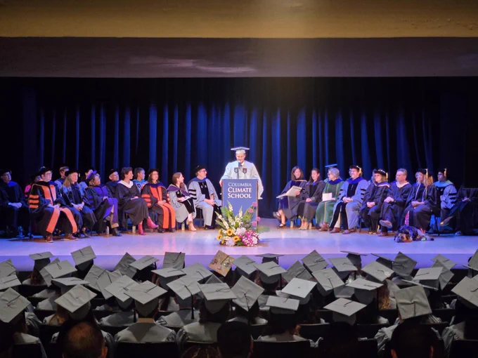 Columbia Climate School faculty and speakers on the stage of Columbia University's Miller Theatre for Class Day 2026, with Evan Moretti, president of Columbia Climate Graduate Council, at the podium and graduates in the audience.