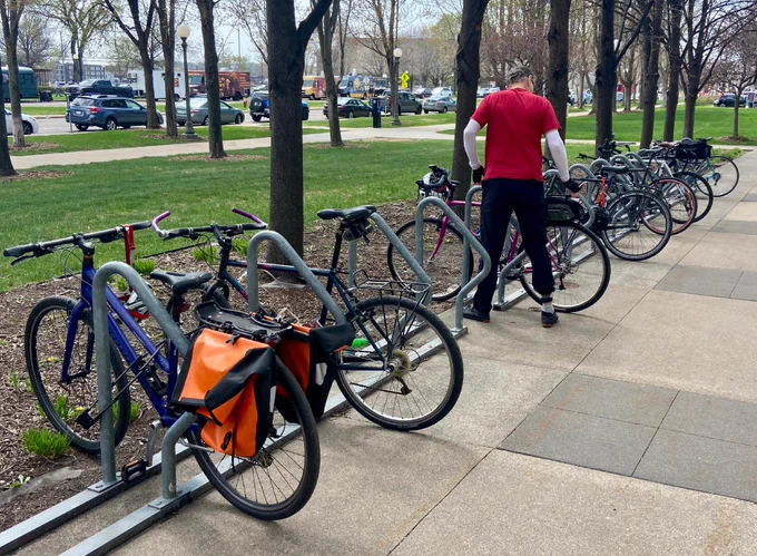 A person locks a bicycle on a bike rack outside of MnDOT’s St. Paul office. Multiple bicycles are secured to the rack. An orange bag is attached to one bike. The background shows John Ireland Boulevard with parked cars and food trucks.