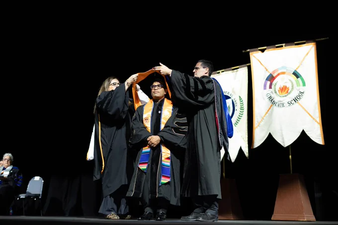 A graduate is receiving a hood during a ceremony on stage, assisted by two faculty members. Behind them are banners, one of which says "Graduate School.” An audience is faintly visible in the darkened background.
