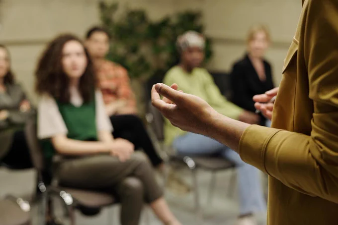 A speaker gestures to a listening seated audience.