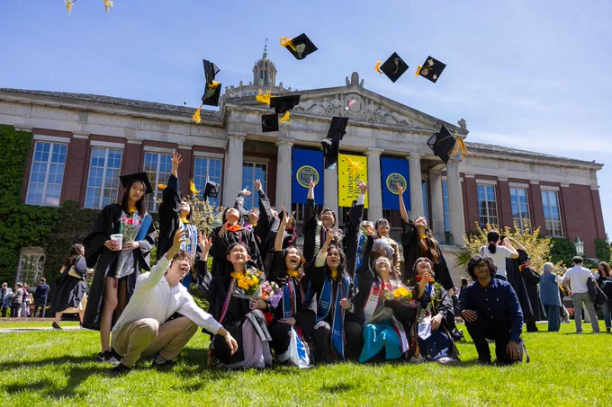 A group of graduates in caps and gowns cheerfully toss their caps into the air in front of Rush Rhees Library. 