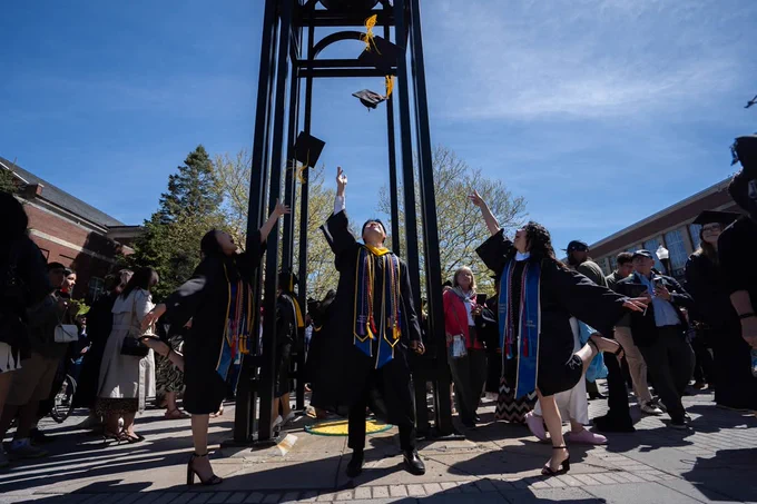 Three students toss their caps in the air in front of the clock tower. 