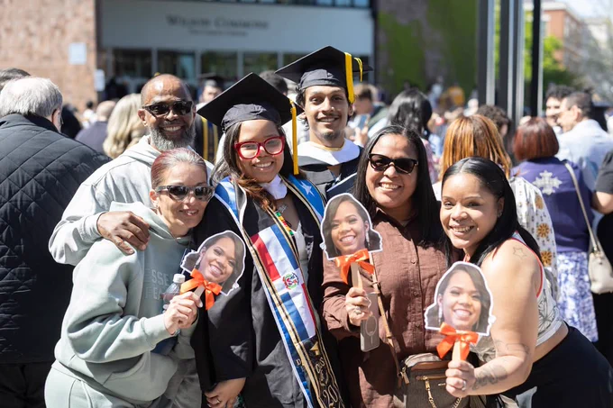 A group of people smiling and posing at a graduation event. Two individuals in the center are wearing graduation caps and gowns. The people around them are holding cutouts of a graduate's face.