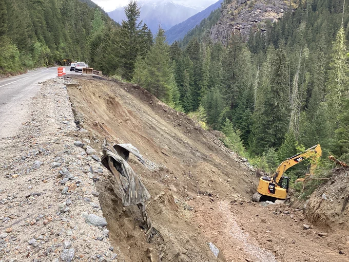 A yellow excavator works on a damaged embankment along State Route 20 in a mountainous area.