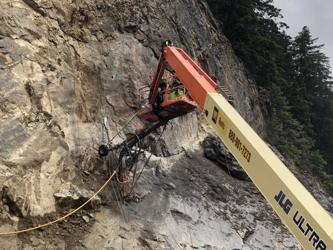 An orange and white supports workers and a drill rig as they work on a steep rock face.