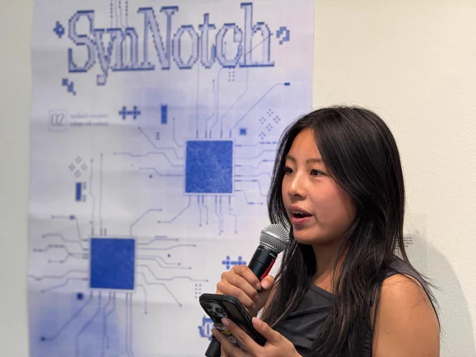 A person is speaking into a microphone while holding a phone. They are standing in front of a poster with the word "SynNotch" and a circuit design in blue.
