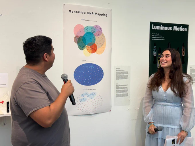 Two people are standing in front of posters on a wall. One holds a microphone and faces a colorful poster titled "Genomics: SNP Mapping." The other person is smiling and looking at the speaker. Another poster titled "Luminous Motion" is partially visible in the background.