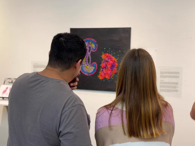 Two people are viewed from behind, looking at a colorful art piece on a wall. The artwork features vibrant colors and abstract shapes on a black background.