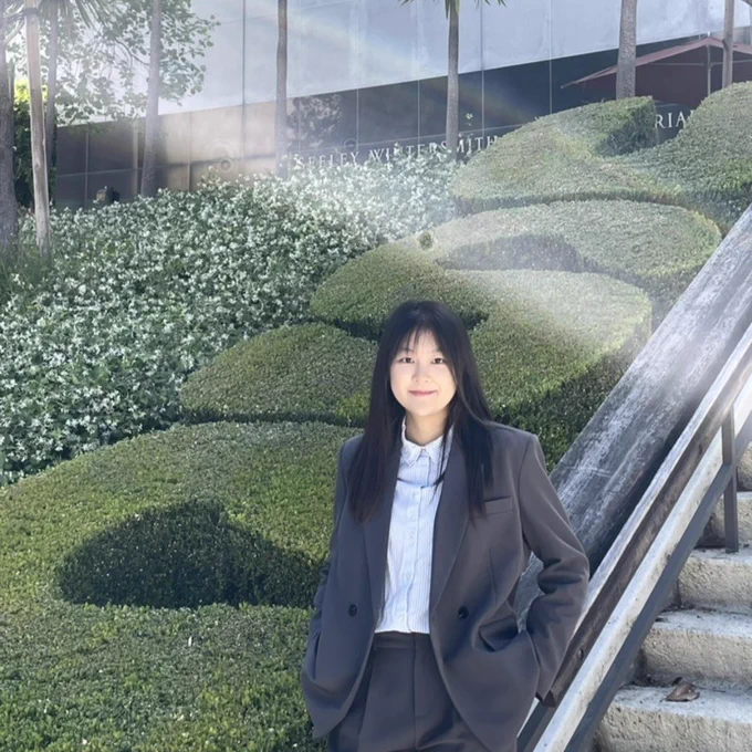 The image shows a person standing outdoors near trimmed shrubs and a staircase. The shrubs are artfully shaped, and there are white flowers in the background.