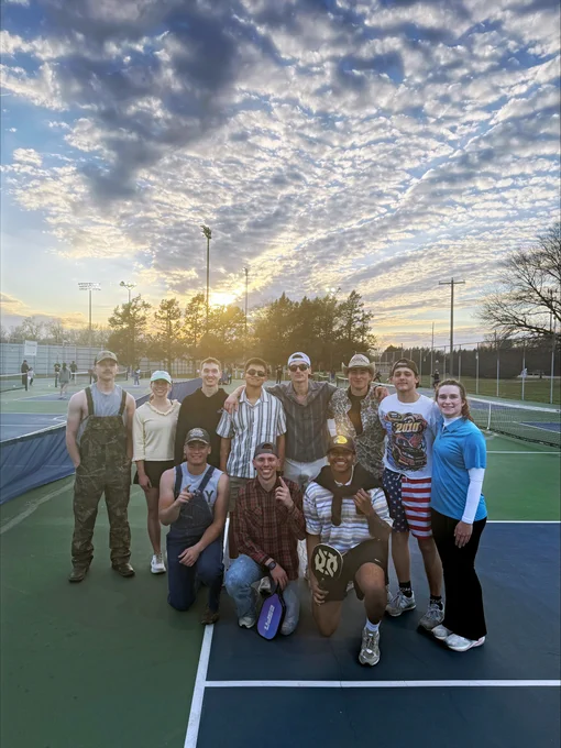 Group of people gathered on a tennis court during sunset. They are casually dressed, with one holding a paddle featuring a Notre Dame logo. The sky is partly cloudy, and trees are visible in the background.