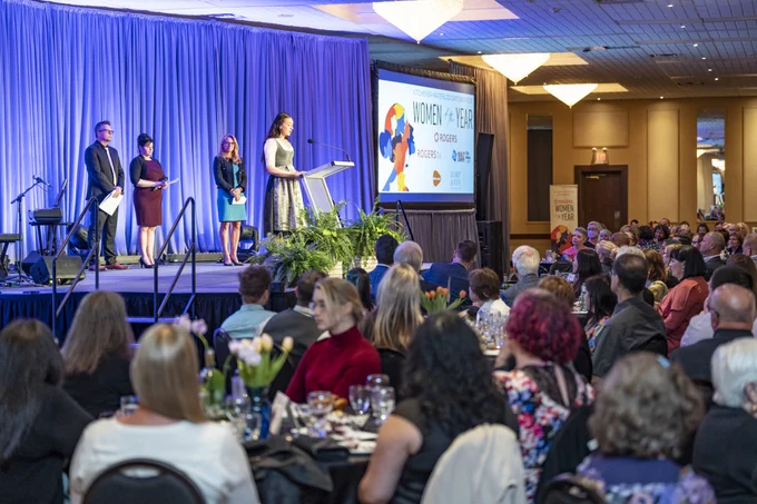 A person is speaking at a podium on a stage with a blue curtain backdrop with a digital screen that reads "Women of the Year." Attendees are seated at tables in the foreground.
