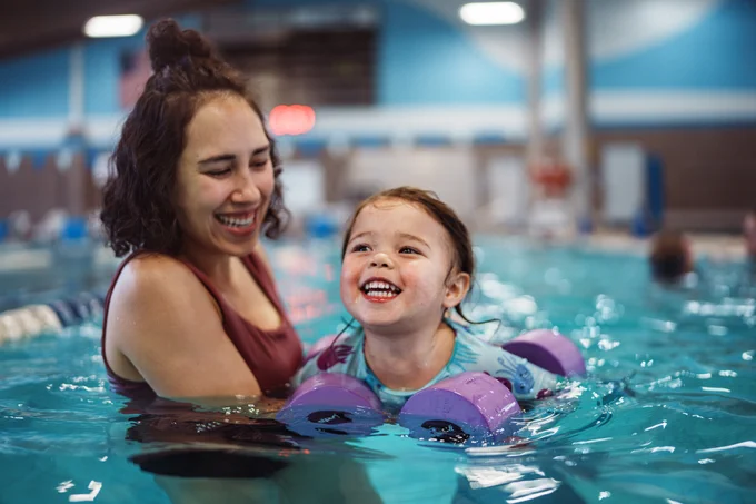 A person and a child are smiling and playing in an indoor swimming pool. The child is wearing purple flotation devices on their arms. The background shows blurred pool equipment and other swimmers.