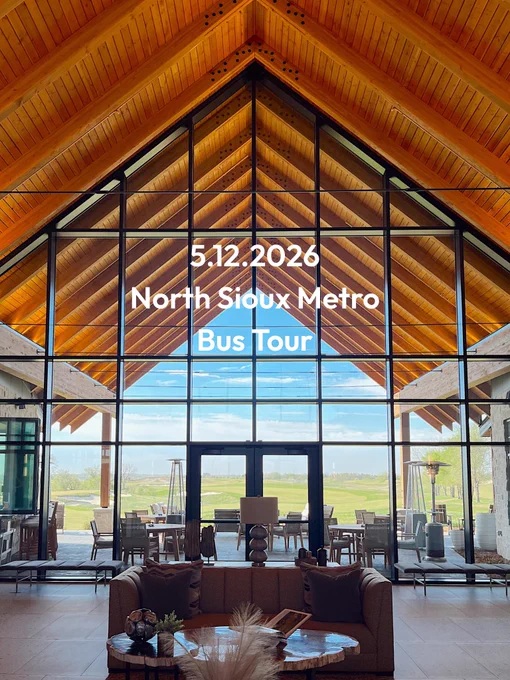 A spacious lobby with large glass windows and a wooden ceiling, offering a view of a green landscape outside. The text displayed reads "5.12.2026 North Sioux Metro Bus Tour." Several chairs and tables are arranged in the lobby.