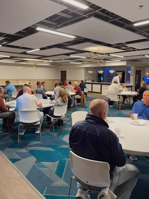 A group of people seated at round tables in a modern room with geometric blue carpeting and a decorative ceiling. A countertop area in the background has a Starbucks Coffee sign.
