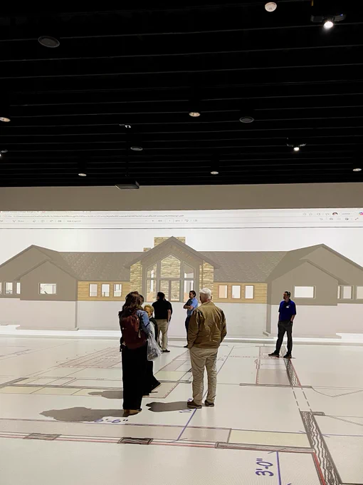 A group of people stand on a large floor map in a dimly lit room, discussing a projected image of a house blueprint displayed on the wall.