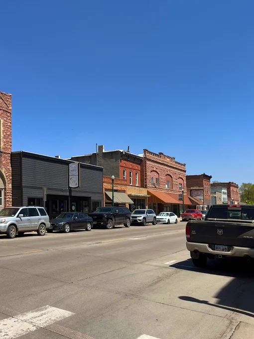 A small-town street scene with parked vehicles lining both sides. On one side, there are several brick and wooden storefronts, each with distinct colors and awnings. The sky is clear and blue.