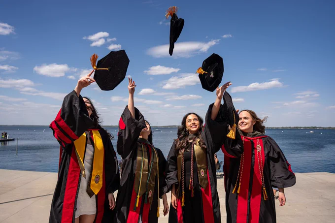 Four graduates in red and black robes celebrate by tossing their caps into the air against a backdrop of blue sky and a large body of water.