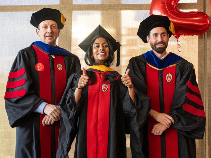 Three individuals in academic regalia stand together, smiling. They are wearing graduation caps and gowns with red and black colors, featuring the University of Wisconsin emblem. 