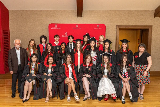 A group of people, some in graduation attire with caps and gowns, and others in formal clothing, stand and sit indoors. The setting looks formal and celebratory.