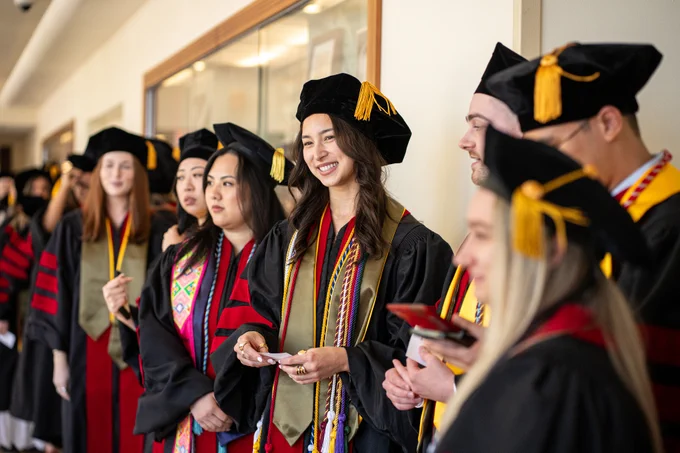 A group of individuals in graduation attire, including caps and gowns with tassels, are standing in a line. They appear to be celebrating, with some holding programs. The setting is indoors, possibly a hallway or corridor. 