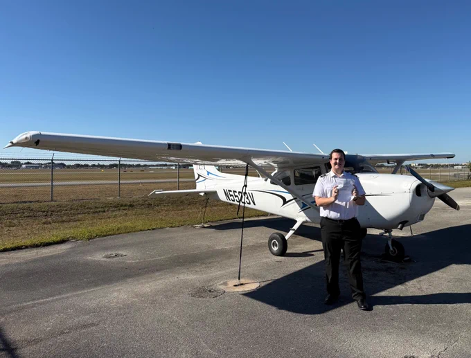 A person in a uniform stands next to a small airplane on an airstrip under a clear blue sky. The aircraft has the registration number N56423V.