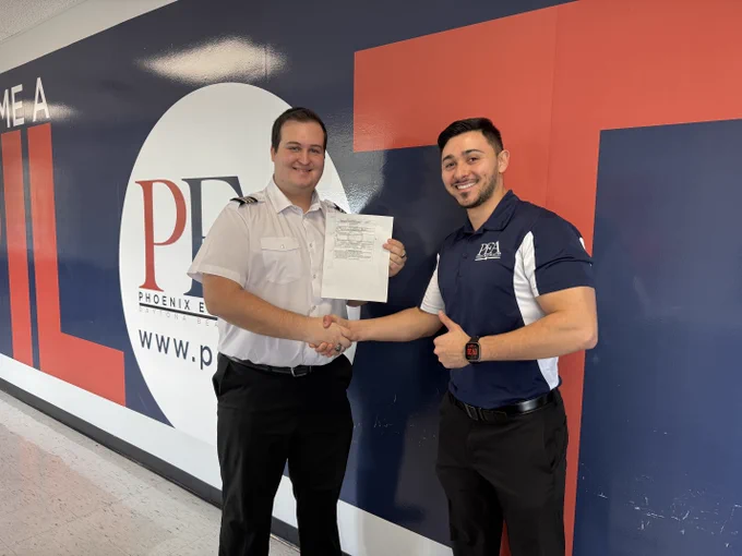 Two people stand smiling inside a building with a large logo for Phoenix East Aviation in the background. One person holds a document, and they shake hands. The environment appears professional and positive.