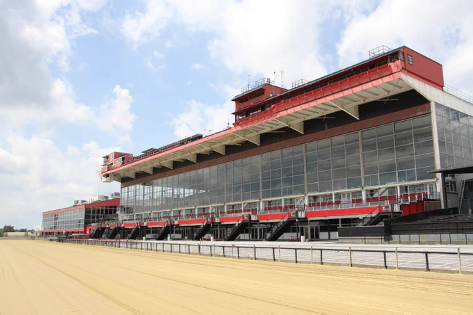 A large, multi-level grandstand at Pimlico Race Course, a horse racing track, with red and white accents, tall glass windows, and a sandy track in the foreground. The scene is under a partly cloudy sky.