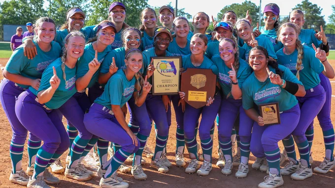 A jubilant FSW softball team poses on the field, wearing teal jerseys and purple pants. They are holding championship plaques and smiling, with many showing number one gestures. The sunlight highlights their faces and vibrant uniforms, while the background shows trees and more people.