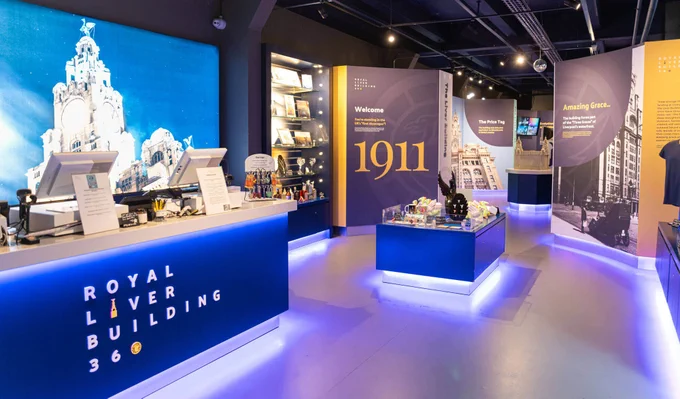 The image shows an interior of a visitor center  at the Royal Liver Building. The room is brightly lit, featuring informational displays about the building's history, including a large "1911" sign. Counters with promotional materials and interactive screens are visible, providing a modern and inviting atmosphere. The walls display images and text relating to the building's significance.