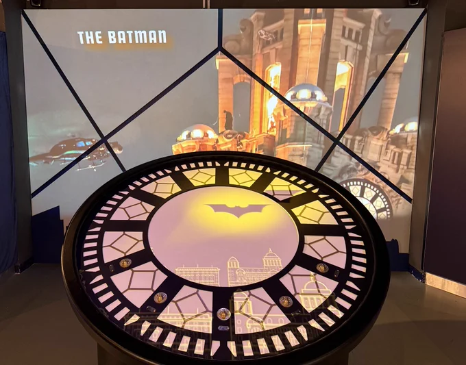 A circular display table with an illuminated Gotham City skyline and Batman symbol in the center. Behind it, a screen shows a scene from "The Batman" featuring a cityscape and futuristic vehicles.