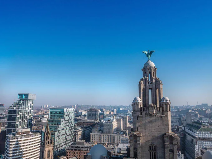 View of Liverpool's skyline featuring the Royal Liver Building with its iconic Liver Bird on top. Surrounding modern and historic buildings are seen under a clear blue sky.