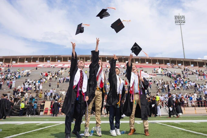 A group of Cornell University graduates in graduation attire celebrates by tossing their caps into the air at Schoellkopf Field at Cornell University. The background shows a large crowd seated in the stadium for a Cornell Commencement ceremony.