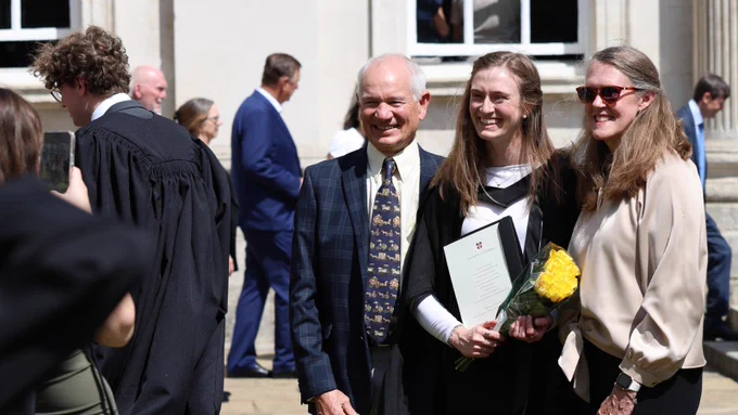 A group of people is posing for a photo outside a building. One person holds a bouquet of yellow flowers and a certificate. Another person is wearing a robe, suggesting a graduation setting. Others in the background are dressed formally.