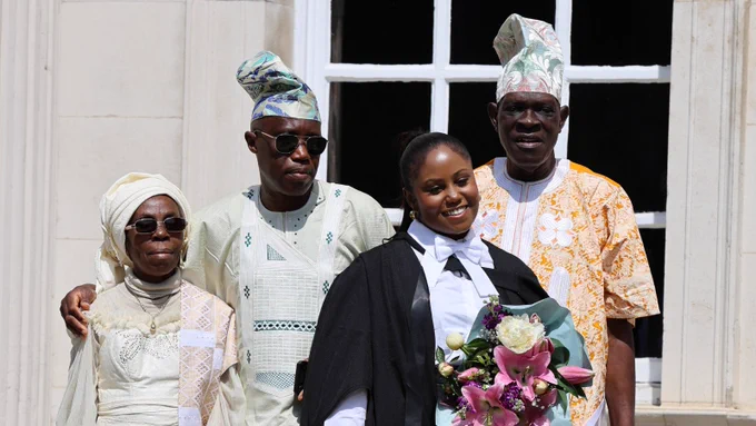 A group photo outside a building with a window. Four people are smiling at the camera. One person in academic attire is holding a bouquet of flowers.