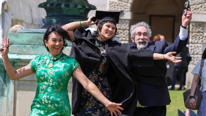 A joyful group celebrates at a graduation. One person is in a cap and gown, flanked by two others in formal attire. They are standing outside, with part of a historic stone building visible in the background.