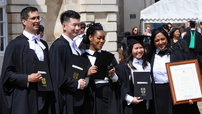 A group of five people dressed in graduation attire, holding diplomas and posing outside a historic building. They appear to be celebrating their achievement.