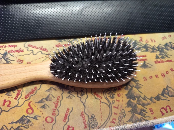 Die Aufnahme zeigt eine ovale Haarbürste mit einem hellen Holzgriff, die flach auf deinem Mittelerde-Mauspad liegt. Der Bürstenkopf ist dicht mit dunklen Naturborsten besetzt, die regelmäßig von feinen, weißen Kunststoffstiften mit abgerundeten Köpfen durchzogen sind. Die Bürste hebt sich durch den hellen Holzton und das tiefe Schwarz der Borsten deutlich von der kartografischen Unterlage ab. Die Kombination aus den weicheren, dunklen Naturborsten und den festeren, hellen Kunststoffstiften verleiht der Bürste eine strukturierte Optik. Im Hintergrund sind auf dem Mauspad die charakteristischen gezeichneten Gebirgszüge und die in Rot gehaltene Schrift von Mittelerde zu sehen. Das Licht betont die natürliche Maserung des Holzes und die detaillierte Anordnung der verschiedenen Borstentypen auf dem Untergrund.