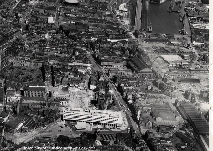 Black and white photograph taken from the air looking over an urban area. A large light building dominates the bottom. A dock is at the top right. There areas where buildings have been cleared and construction appears to be in progress