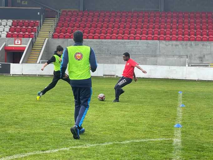 A photo of a football pitch with players from two tams running and tackling each other. One team is dressed in red and the other is in neon yellow vests. 