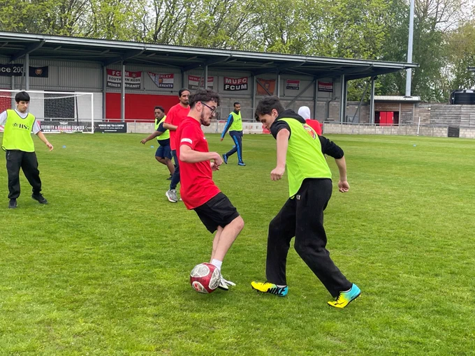 A photo of a football pitch with players from two tams running and tackling each other. One team is dressed in red and the other is in neon yellow vests. 